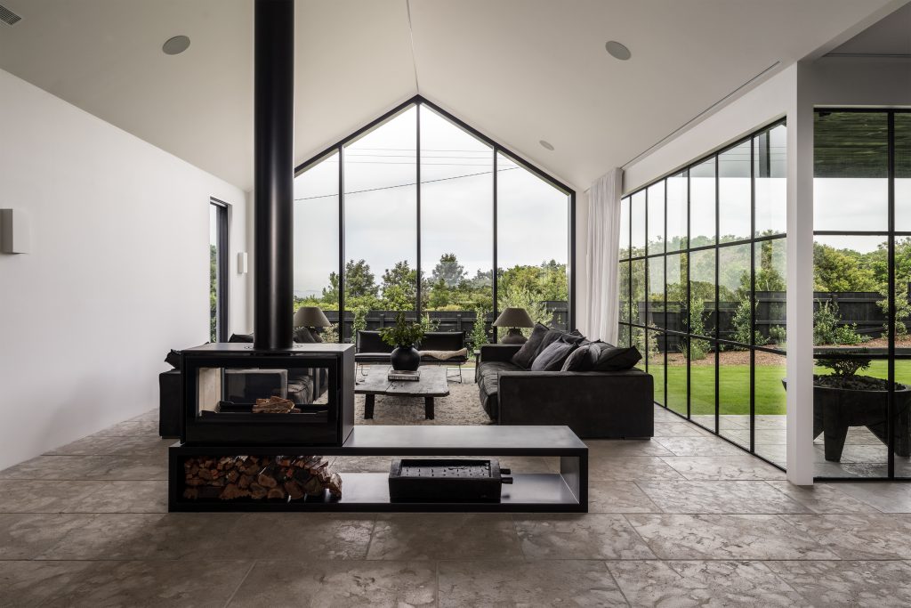 Nox Byron Bay living room with fireplace, stone tiled floor, high pitched ceiling and large glass windows